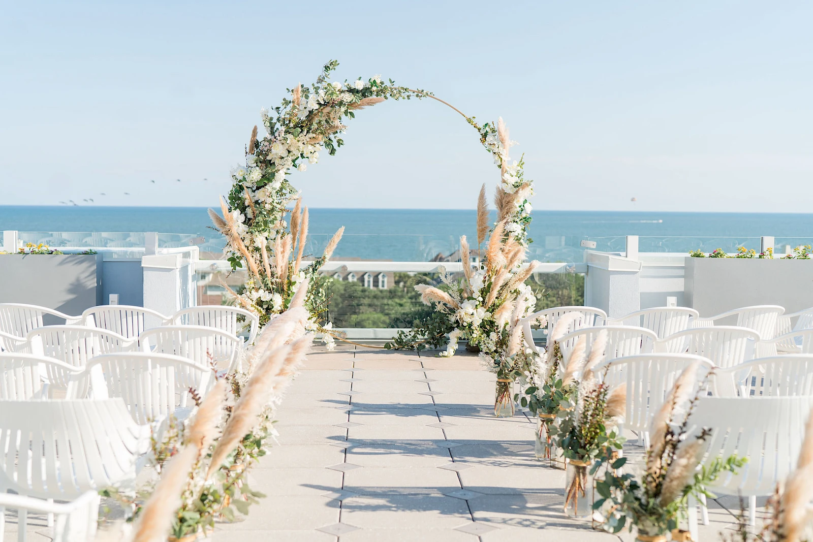 A beachside wedding setup with a floral archway over an aisle, white chairs along a tiled path, and the ocean in the background.