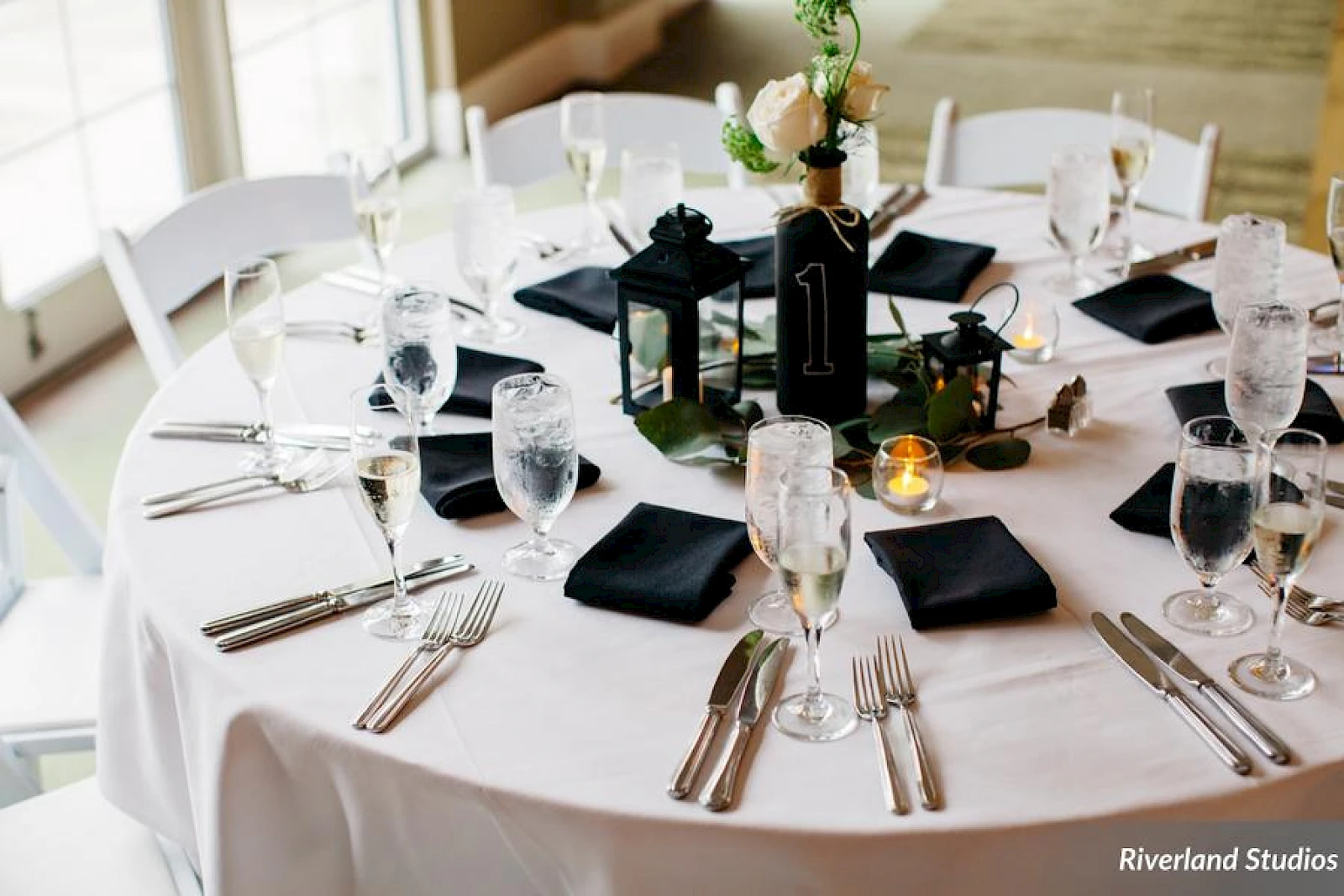 A round table set for a formal event with white tablecloths, glassware, silverware, black napkins, candles, and a centerpiece; elegant dining setup.
