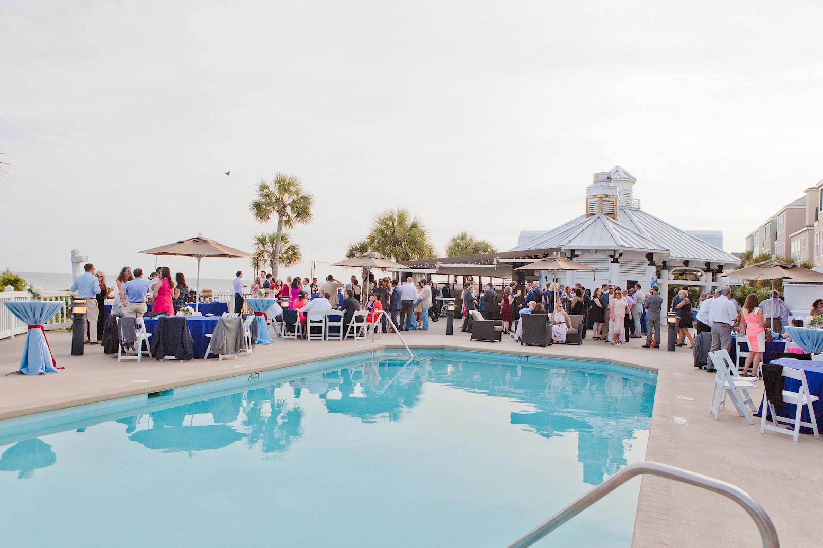 A lively poolside party with groups of guests mingling near a blue swimming pool, white tables, umbrellas, and a white vendor tent in the background.