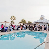 A lively poolside party with groups of guests mingling near a blue swimming pool, white tables, umbrellas, and a white vendor tent in the background.