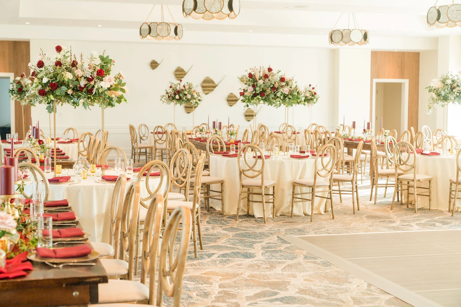 A spacious banquet hall set for a formal event with round tables, cream tablecloths, red napkins, elegant chairs, and tall floral centerpieces.