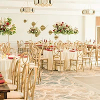 A spacious banquet hall set for a formal event with round tables, cream tablecloths, red napkins, elegant chairs, and tall floral centerpieces.