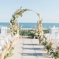 An outdoor wedding ceremony setup by the sea with a floral arch, white chairs, and a sunny, clear sky, perfect for beach vows.