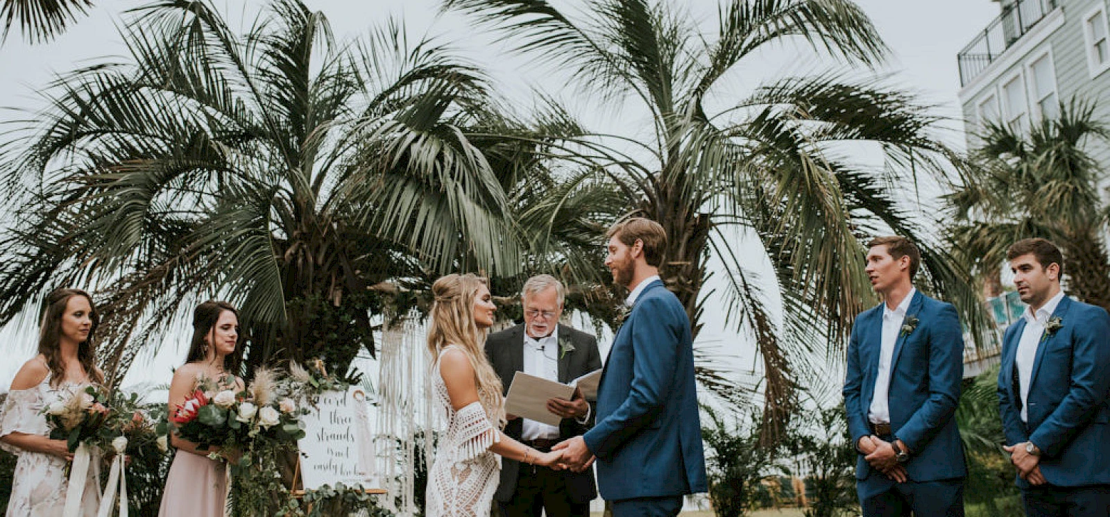 A wedding ceremony outdoors with a bride and groom exchanging vows, officiant, bridesmaids holding bouquets, and groomsmen in blue suits under palm trees.
