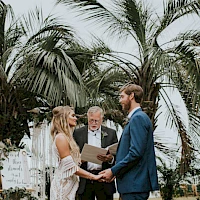 A wedding ceremony outdoors with a bride and groom exchanging vows, officiant, bridesmaids holding bouquets, and groomsmen in blue suits under palm trees.