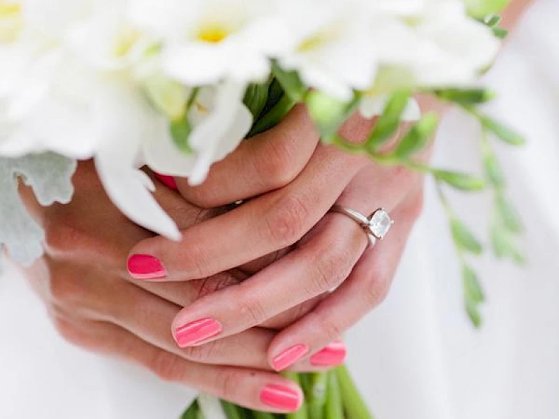 A bouquet of white flowers being held by someone with pink nails and a ring, wrapped stems visible.