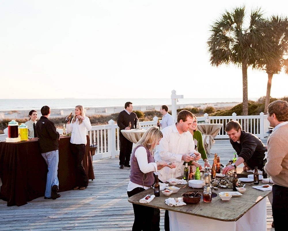 A beachside outdoor party with guests mingling and a buffet set up on a wooden deck near the ocean at sunset.