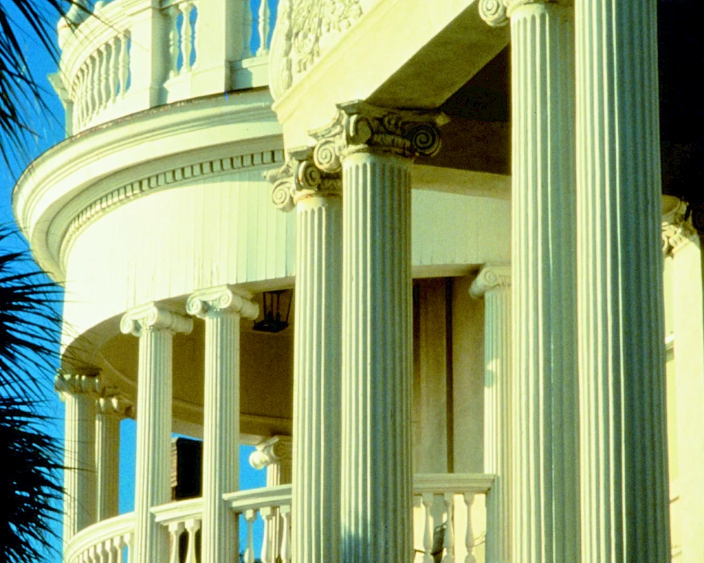 A grand, cream-colored balcony with tall fluted columns, ornate railing, and a curved balcony overlook against a bright blue sky, luxury style architecture.