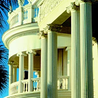 A grand, cream-colored balcony with tall fluted columns, ornate railing, and a curved balcony overlook against a bright blue sky, luxury style architecture.