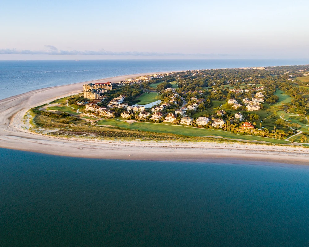Aerial view of a crescent beach with turquoise water, a sandy shoreline, and a cluster of green, villa-style houses along the coast.