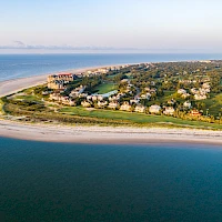 Aerial view of a crescent beach with turquoise water, a sandy shoreline, and a cluster of green, villa-style houses along the coast.