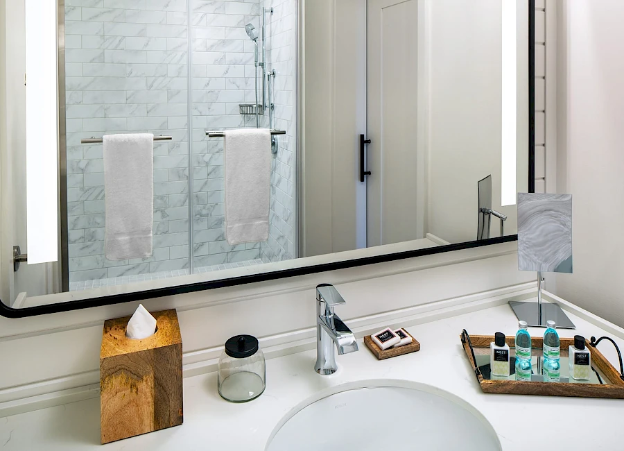 A modern bathroom vanity with a large mirror, marble-tiled shower, and a clutter-free counter featuring tissues, soap, and a wooden tray with amenities, all under bright lighting.