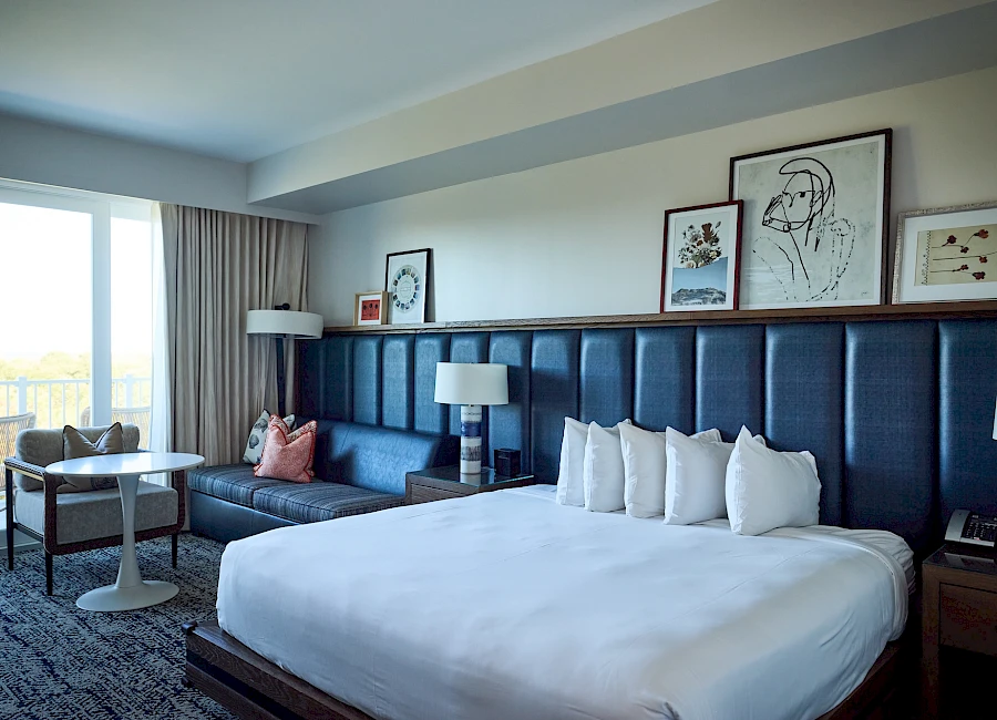 A modern hotel room with a large bed, blue padded headboard, nightstands, lamps, seating area, framed art above the bed, and a balcony view.