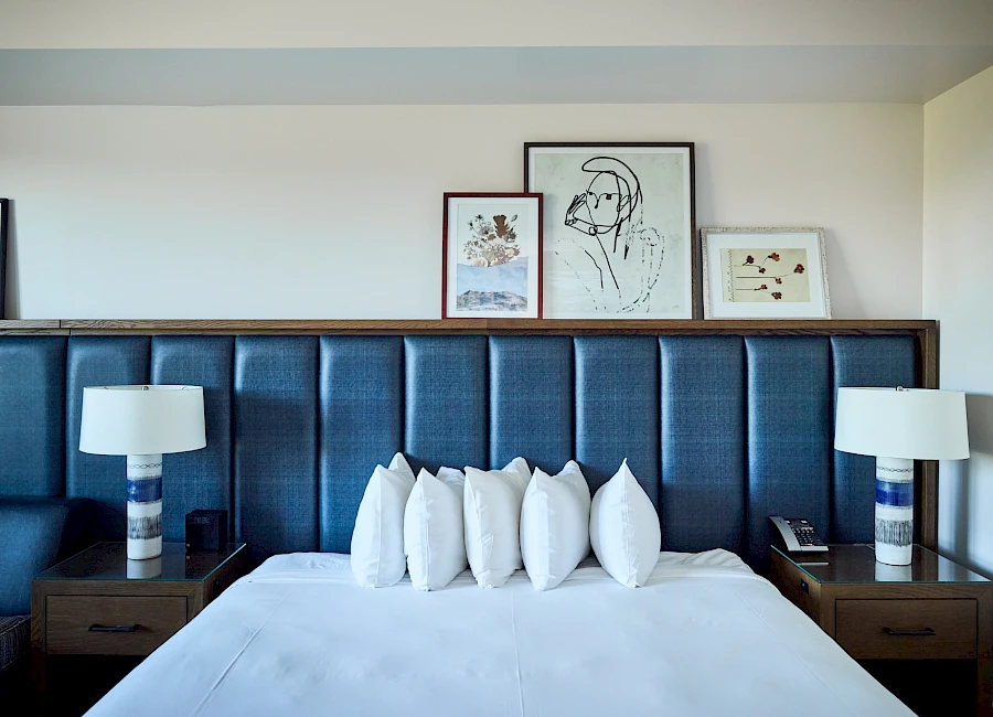 A modern hotel bedroom with a large upholstered blue headboard, white bedding and pillows, two matching nightstands with lamps, and framed art above the bed, ending with a neat, clean look.
