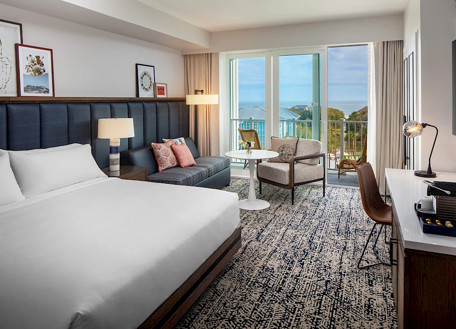 A modern hotel room with a king bed, a sofa, two chairs by a balcony with ocean view, a desk, artwork, lamps, and a patterned carpet.