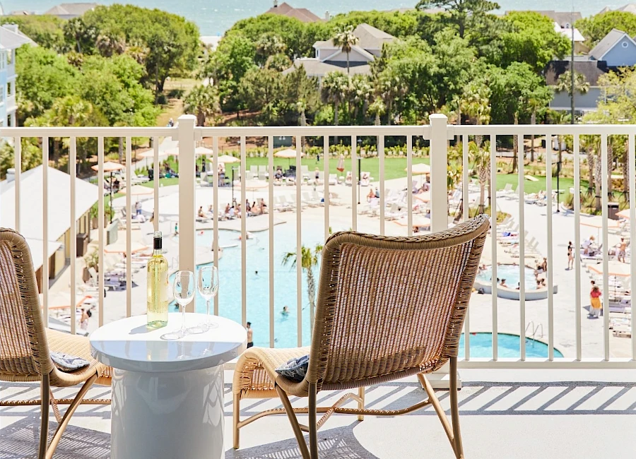 A balcony view overlooking a resort pool with lounge chairs, greenery, and the ocean in the distance; two chairs and a small table frame the scene.