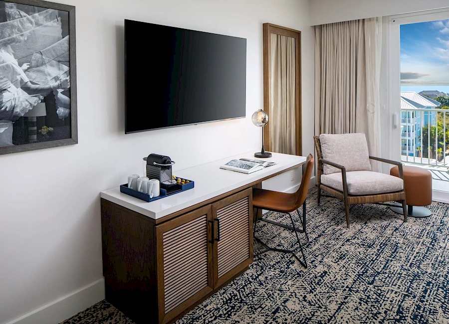A modern hotel room with a wall-mounted TV, a desk with a chair, a small cabinet, a bedside lamp, and a seating area near a balcony window.