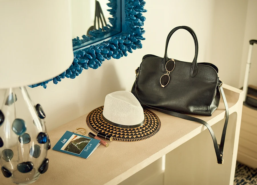 A stylish bedside setup with a black handbag, a white hat, a patterned coaster, a blue-bordered mirror, a lamp with a dotted vase, and a small card on a cream table.