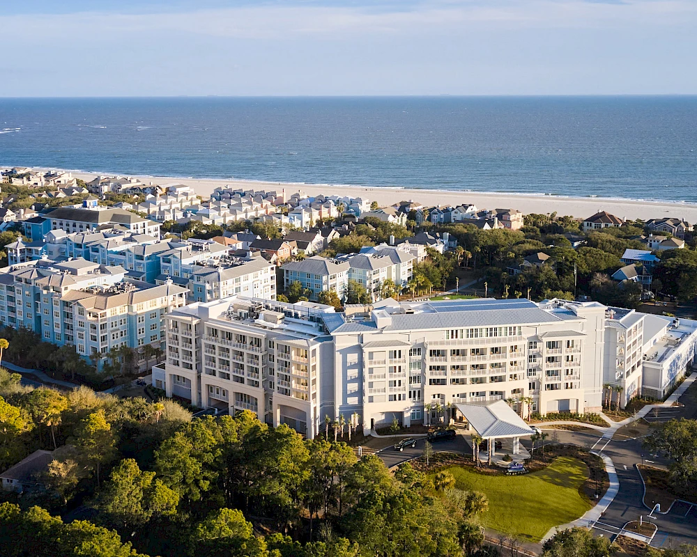 A coastal resort town with white apartment buildings near a sandy beach and blue ocean, surrounded by green trees and hills.