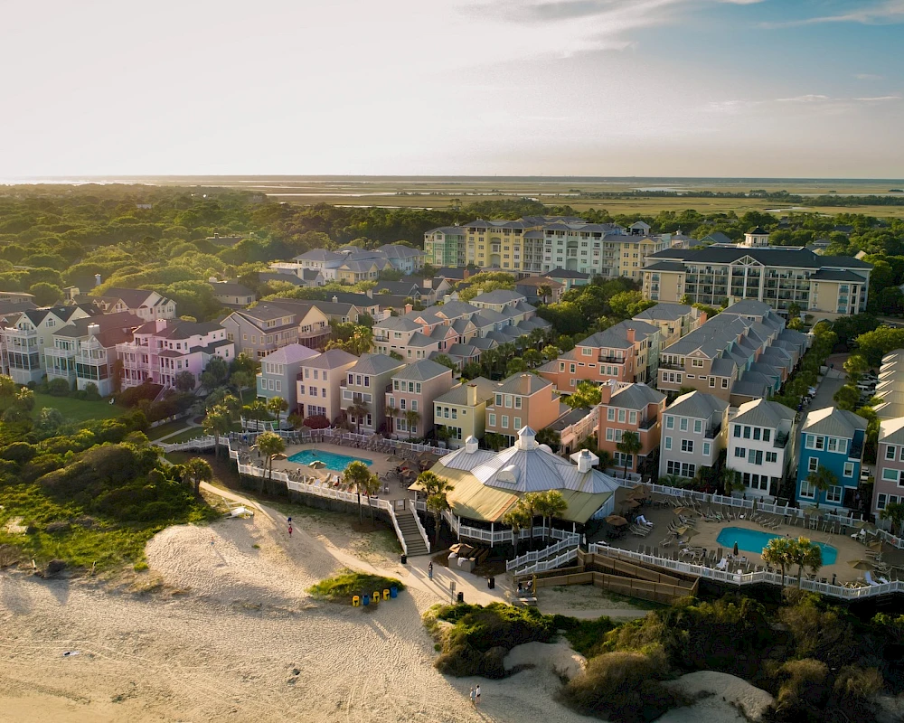 Aerial view of a coastal resort with colorful houses along the shore, beaches, and a few pools in the foreground, sunny and serene.