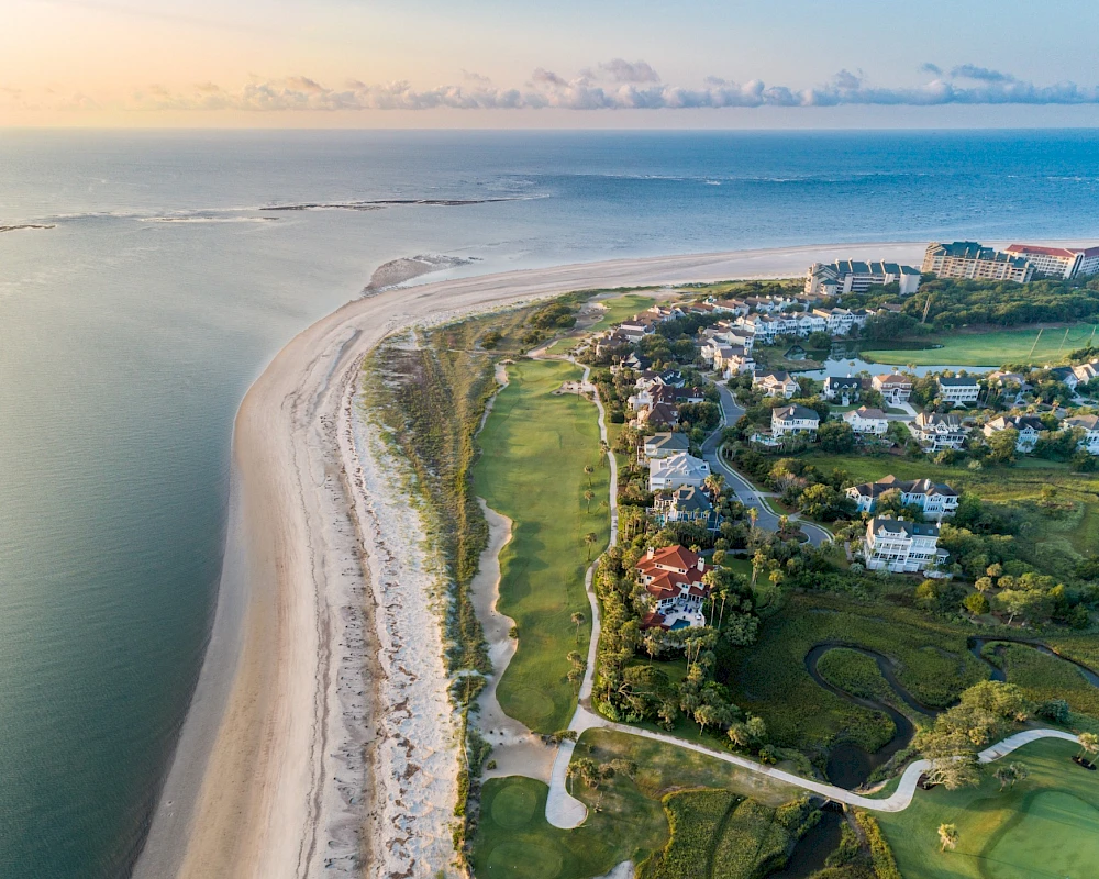 Aerial view of a coastal town with a long sandy beach, calm ocean, and green lawns; houses line the shore and a winding path runs through.
