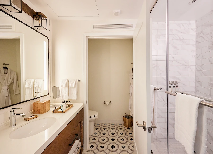 A modern hotel bathroom with a double sink vanity, framed mirror, marble shower, patterned floor, and a separate toilet room in the back.