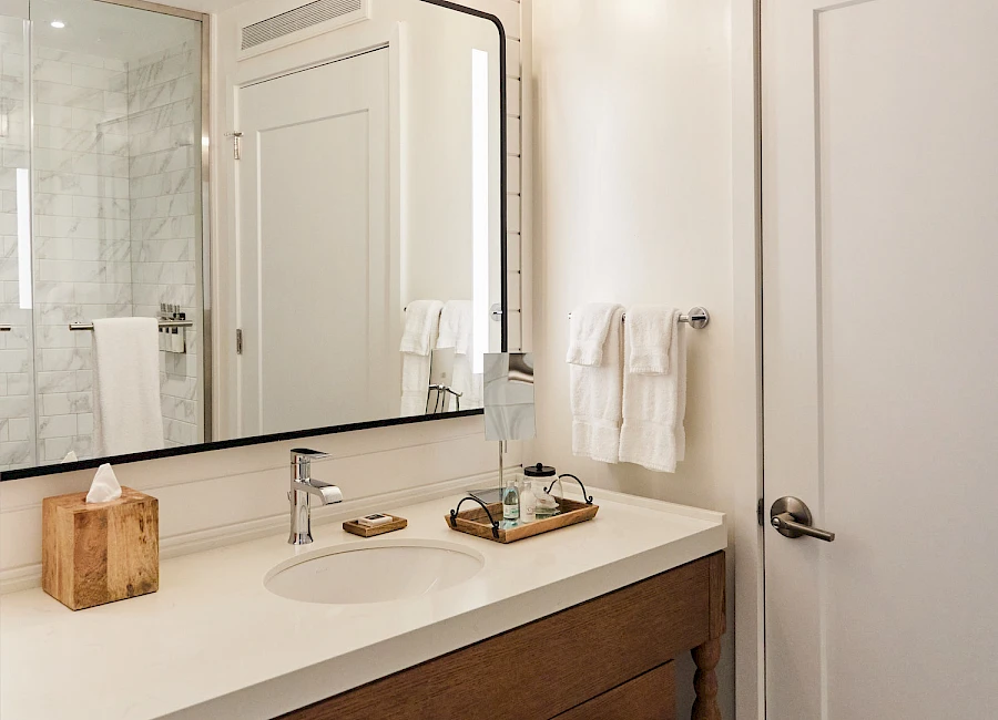 A stylish bathroom with a large framed mirror, warm lighting, a wood vanity, white countertop, two sinks, towels, and a glass shower in the background.