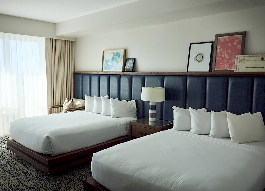 A hotel room with two large beds, a dark upholstered headboard, lamps, framed art, and a large window with sheer curtains.