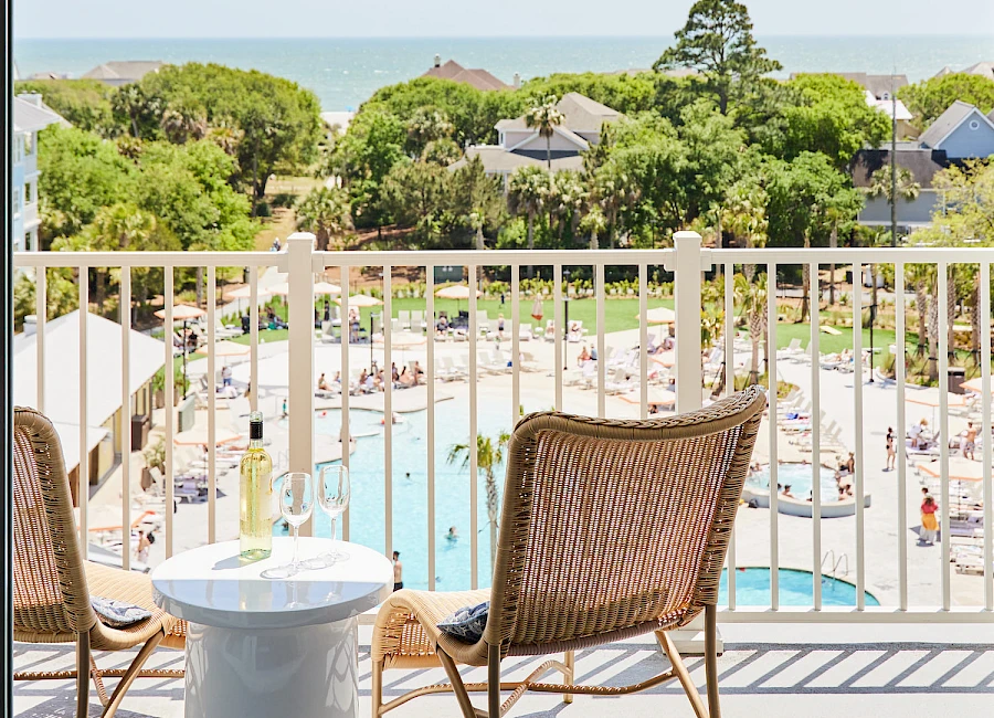 A sunny resort balcony with two chairs and a small table overlooking a pool, greenery, and the ocean in the distance.