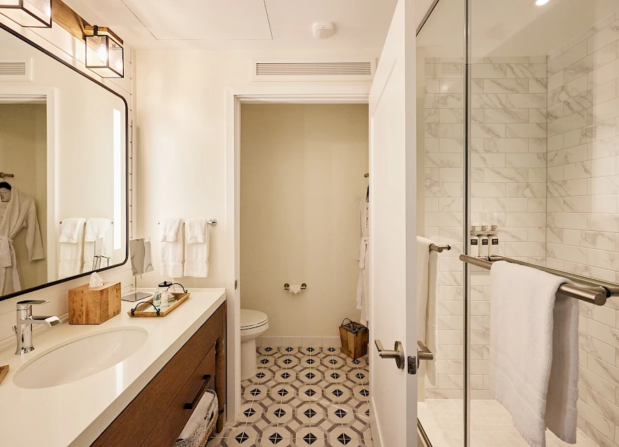 A modern bathroom with a double-sink vanity, large mirror, glass shower, white marble accents, patterned tile floor, and robes by the doorway, neat and bright.