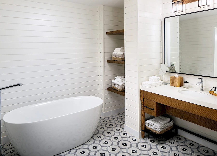 A modern bathroom featuring a freestanding white tub, wooden vanity with sink, open shelving, patterned tile floor, and a large mirror.