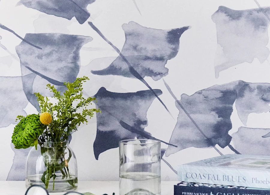A tidy desk with a glass vase of greenery, a few yellow flowers, a glass cup, reading glasses, a stack of books, and a soft blue-gray abstract wallpaper.