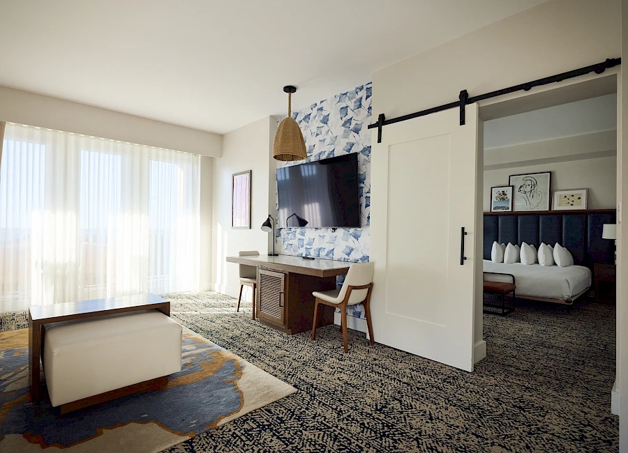 A modern hotel suite with a living area and an open doorway to a bedroom; features a wall-mounted TV, decorative blue wallpaper, sliding barn door, and a desk.