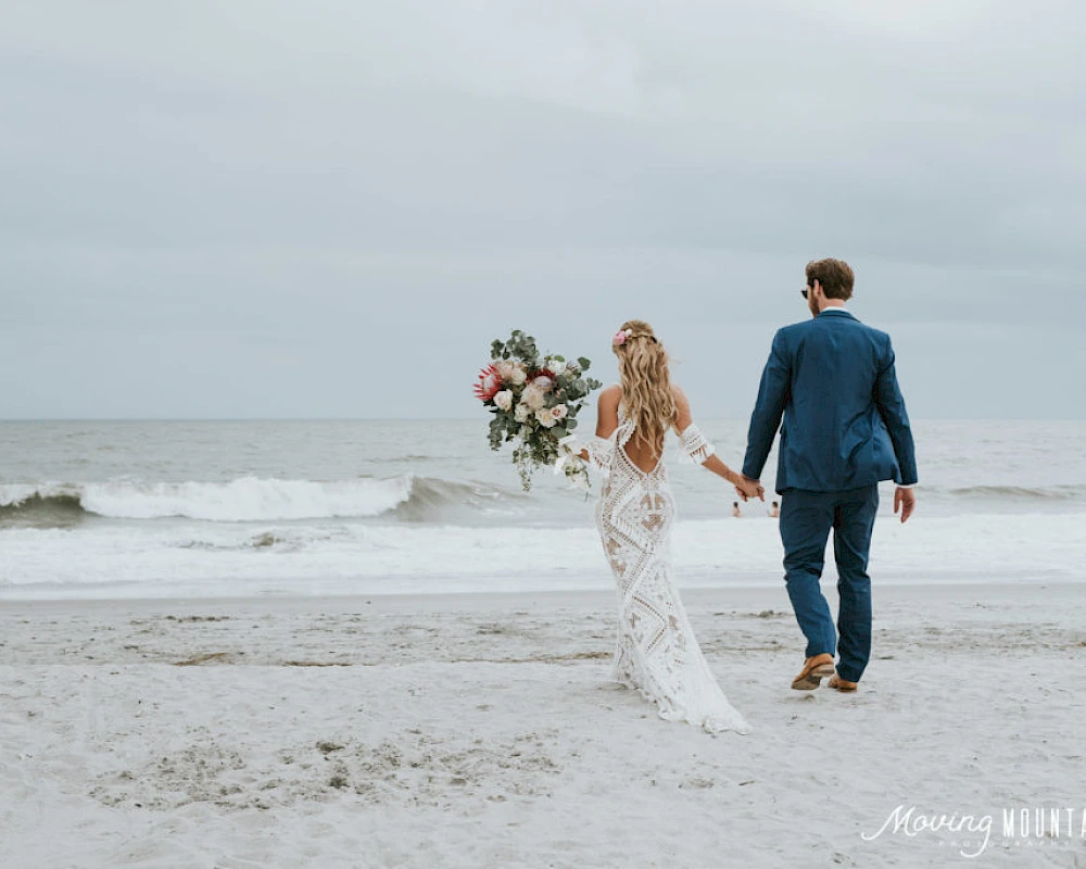 A bride in a white lace dress and a groom in a blue suit walk hand-in-hand along a sandy beach, with the bride holding a bouquet, waves in the background.