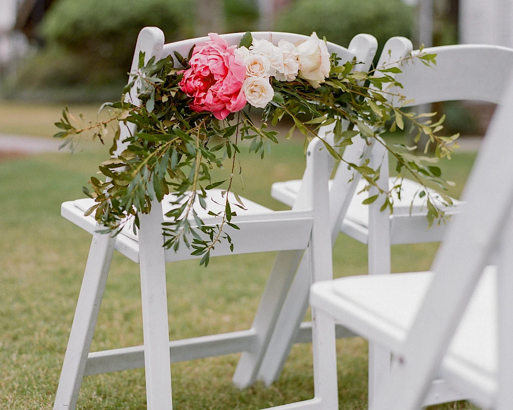 White folding chairs line a grassy aisle, adorned with a delicate floral garland of pink and white blooms and greenery.