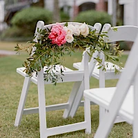 White folding chairs line a grassy aisle, adorned with a delicate floral garland of pink and white blooms and greenery.