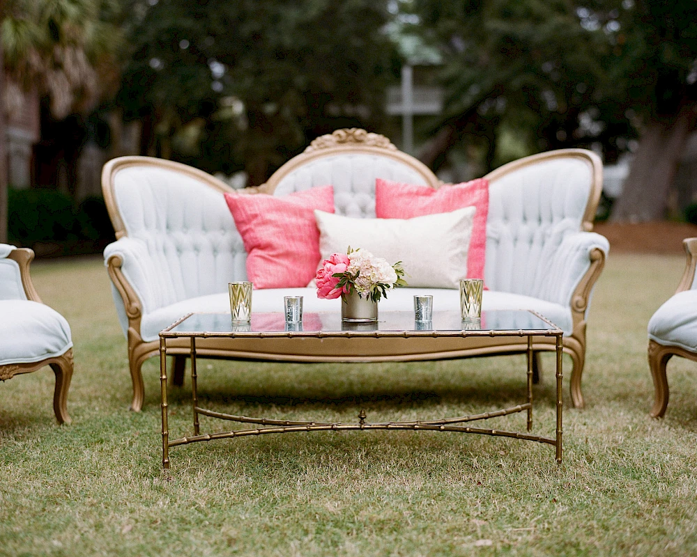 A stylish outdoor seating set with white tufted sofas, pink cushions, and a gold coffee table on a grassy lawn, perfect for a chic garden gathering.