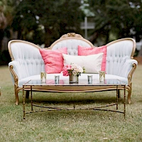A stylish outdoor seating set with white tufted sofas, pink cushions, and a gold coffee table on a grassy lawn, perfect for a chic garden gathering.