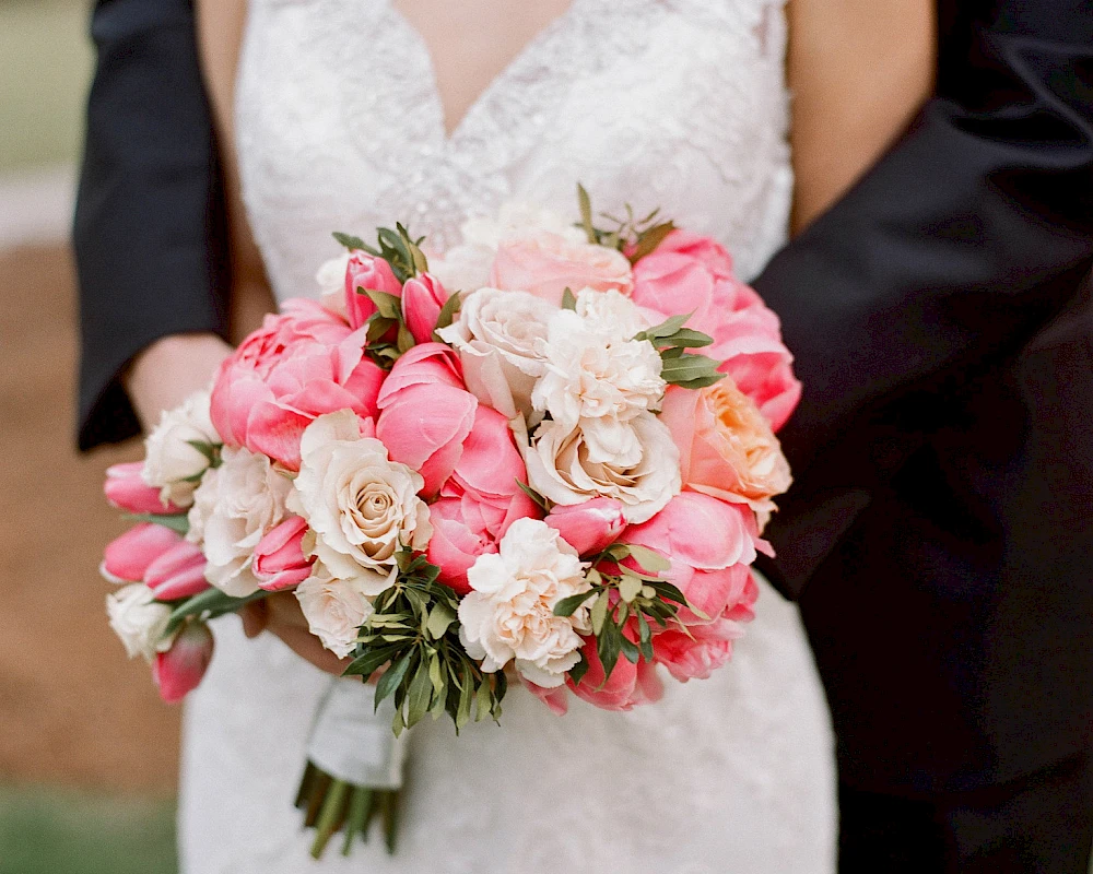 A bride holds a bouquet of pink and white roses and peonies while wearing a lace wedding dress, beside a groom in a suit. End.