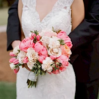 A bride holds a bouquet of pink and white roses and peonies while wearing a lace wedding dress, beside a groom in a suit. End.