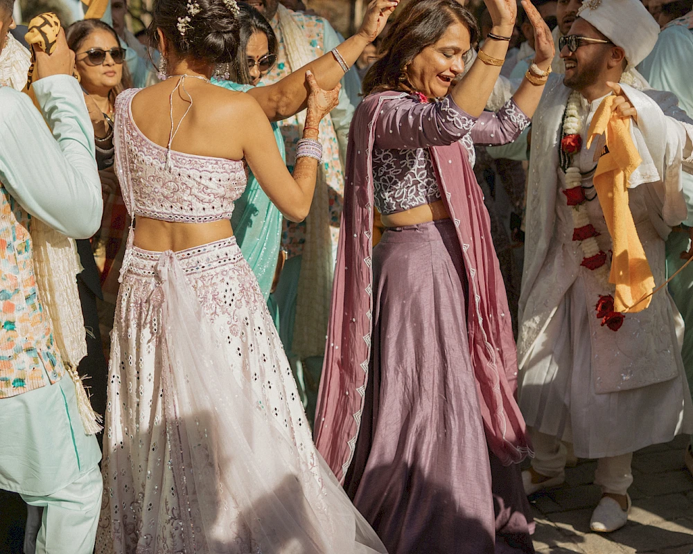 People dancing joyfully in colorful traditional outfits at an outdoor celebration, surrounded by onlookers and trees, sunny day vibes.