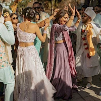 People dancing joyfully in colorful traditional outfits at an outdoor celebration, surrounded by onlookers and trees, sunny day vibes.