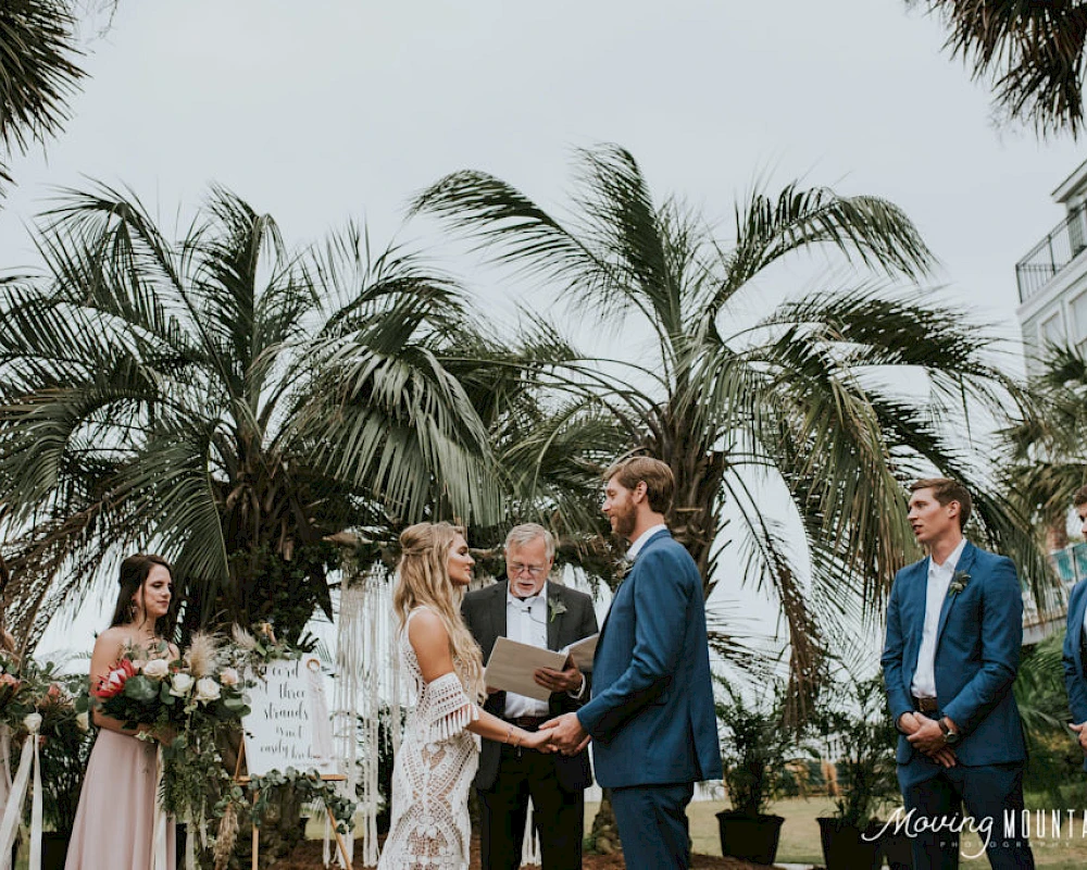 A wedding ceremony outdoors with a bride and groom exchanging vows, bridesmaids and groomsmen standing, tropical palm backdrop, officiant reading vows.