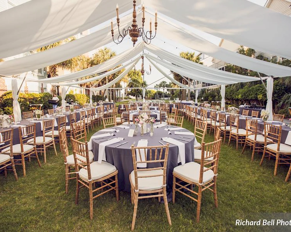 Outdoor wedding reception setup with round tables, blue tablecloths, gold chairs, draped white fabric canopy, chandeliers, and a grassy venue.