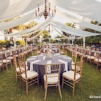 Outdoor wedding reception setup with round tables, blue tablecloths, gold chairs, draped white fabric canopy, chandeliers, and a grassy venue.