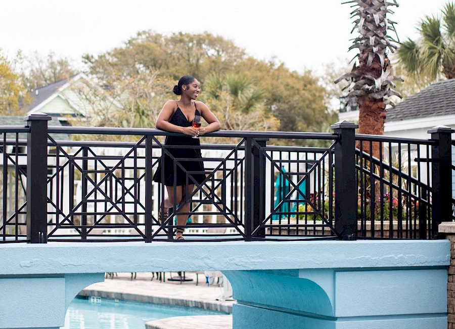 A person in a black outfit stands on a decorative bridge over a turquoise pool, palm trees and tropical scenery in the background.