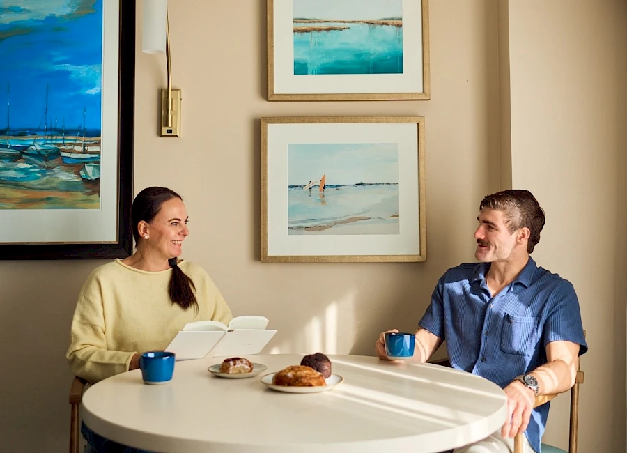 Two people sit at a round table chatting over coffee and pastries in a cozy, art-filled room with framed seascapes on the wall.