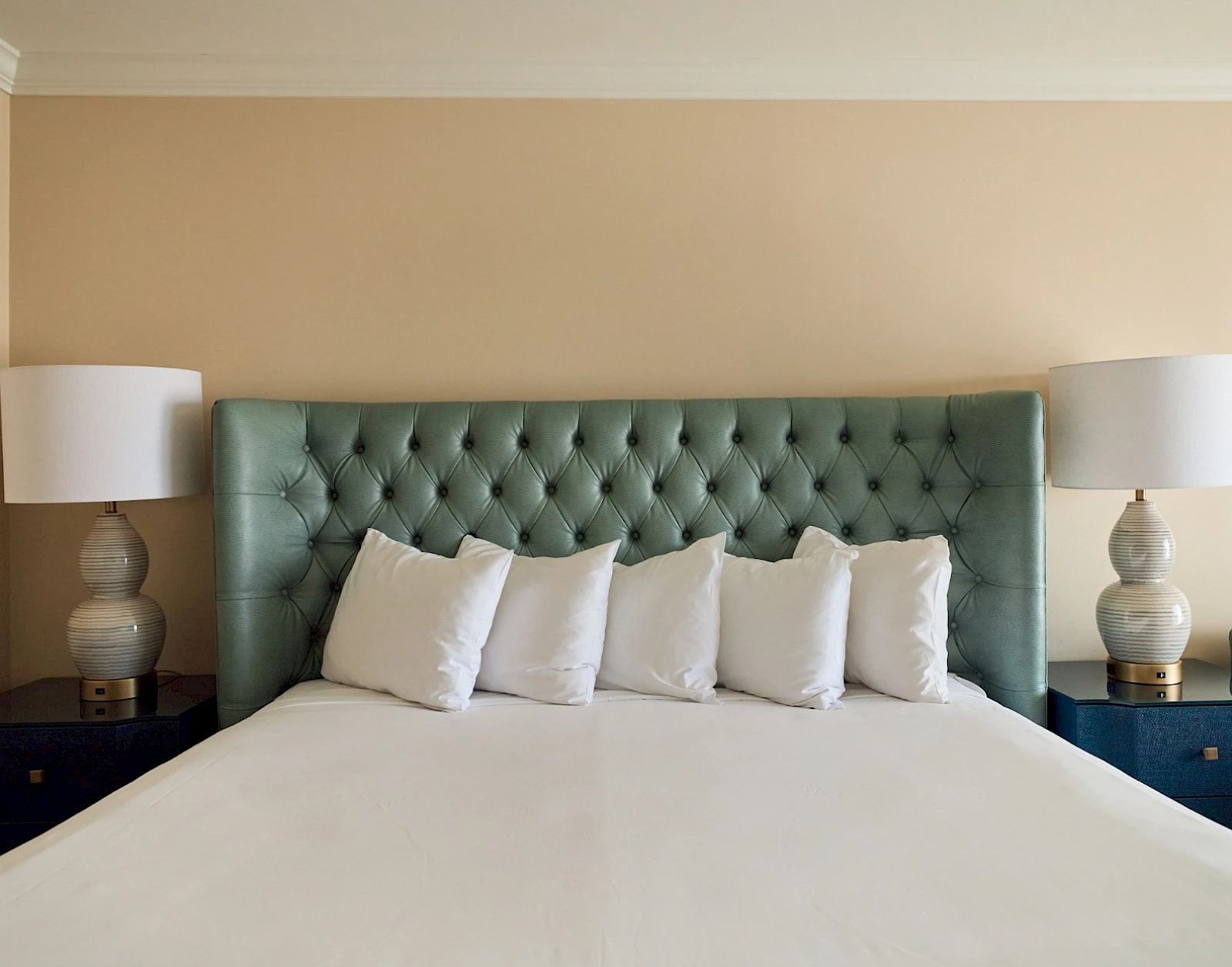 A neatly made bed with a teal tufted headboard, white bedding, and five white pillows flanked by matching nightstands with lamps.