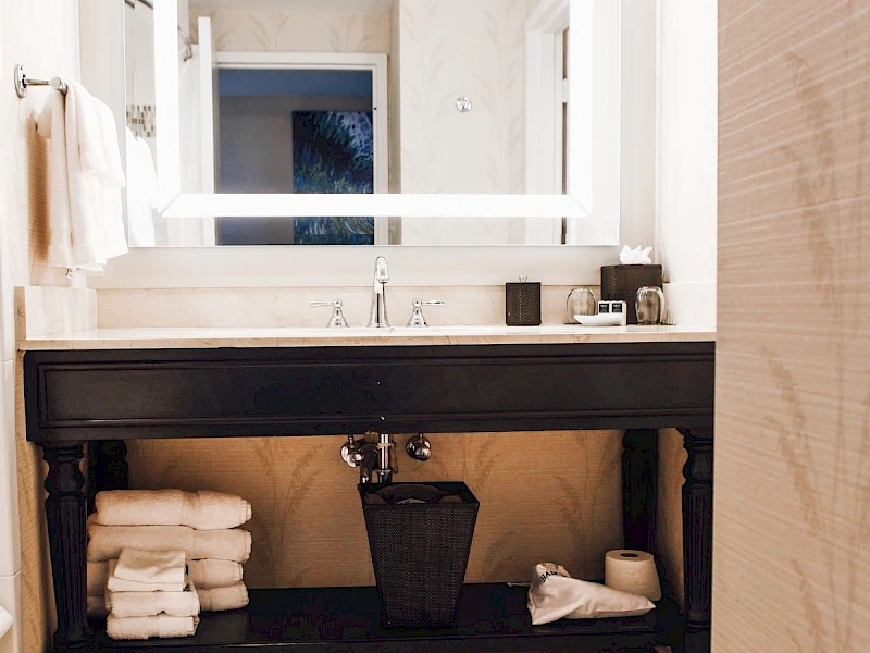 A modern bathroom with a dark wooden vanity, white illuminated mirror, stacked towels, a small trash bin, and a cozy, clean vibe.