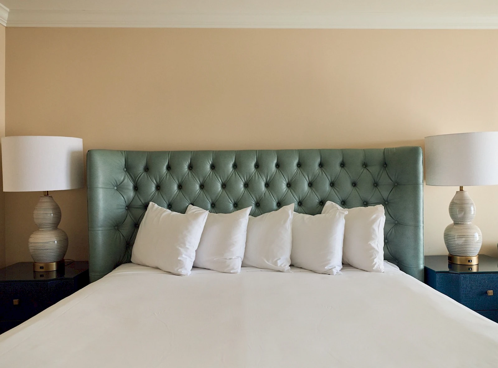 A neatly made bed with a tufted green headboard, five white pillows, matching nightstands, and two lamps flanking the bed.